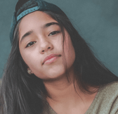 A teenage girl of South Asian ethnicity wears a backward baseball cap and looks directly at the camera with a neutral, calm expression.