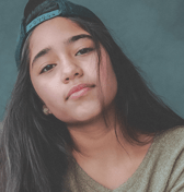 A close-up portrait of a teenage girl with long dark hair, wearing a backwards baseball cap and a light-colored sweater. She looks directly at the viewer with a neutral and thoughtful expression.