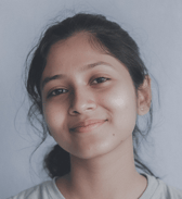 A smiling teenage girl of South Asian ethnicity looks directly at the camera with a gentle expression against a light, plain background.