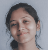 A young teen girl with dark hair tied back smiles gently while looking directly at the viewer, set against a plain, light background.