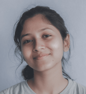 A smiling teenage girl with dark hair pulled back looks directly at the camera, conveying a calm and approachable mood. She is in an indoor setting against a plain light background.
