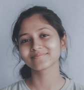 A young South Asian woman, likely a teen, smiles gently while looking directly at the viewer. She has dark hair pulled back and wears a light t-shirt against a plain background, projecting a calm and friendly demeanor.