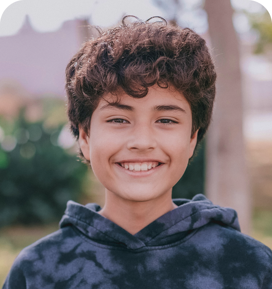 A smiling child with short, curly brown hair, wearing a dark tie-dye hoodie, looks directly at the camera. The background is a softly blurred outdoor setting with trees and greenery, suggesting a bright and positive mood.