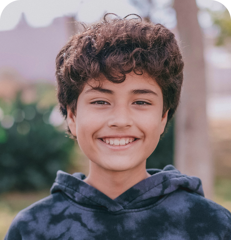 A young boy with dark curly hair smiles confidently at the camera. He is wearing a tie-dye hoodie and is outdoors with a softly blurred natural background.