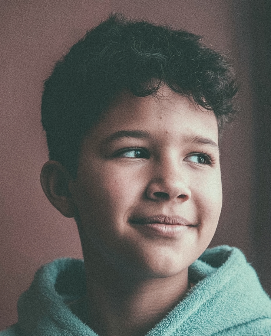 A smiling Hispanic child with curly hair looks thoughtfully to the side while wearing a comfortable hooded garment. The close-up portrait conveys a calm and hopeful mood.