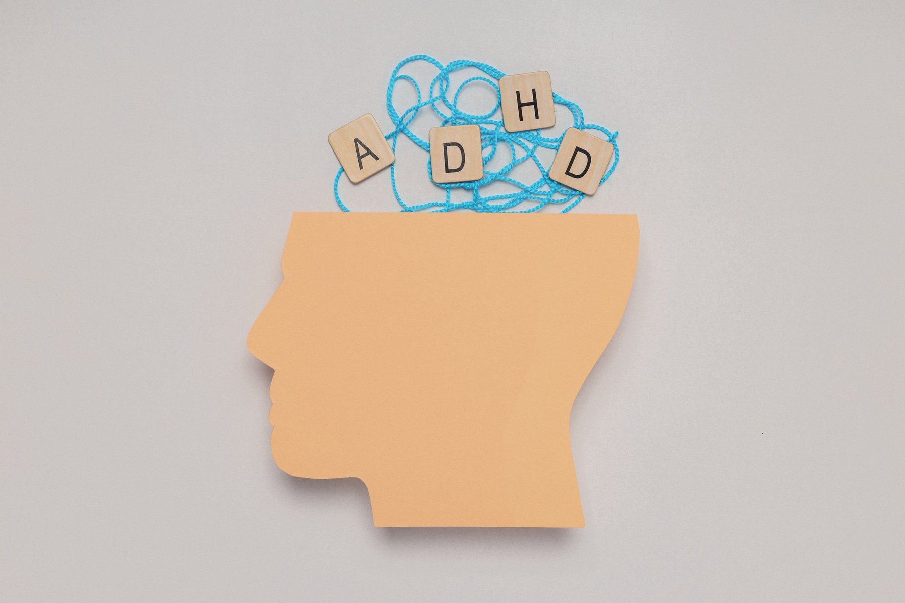 A light brown paper cutout of a human head in profile with tangled blue string and wooden letter tiles spelling 'ADHD' appearing above it. This conceptual image represents the mental health condition of ADHD.