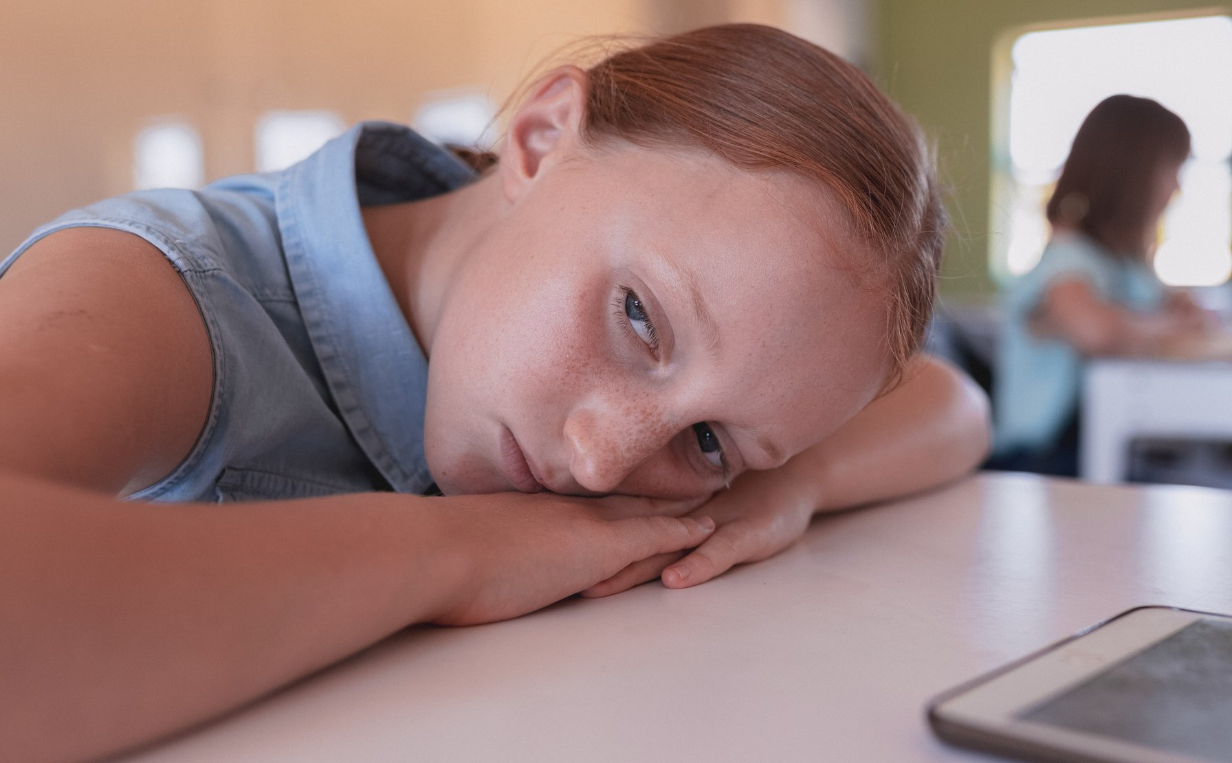 A young girl with reddish-brown hair rests her head on her arms on a table, looking directly at the viewer with a subdued and thoughtful expression. She is in an indoor setting, possibly a classroom, with a blurred figure seated at a desk in the background.