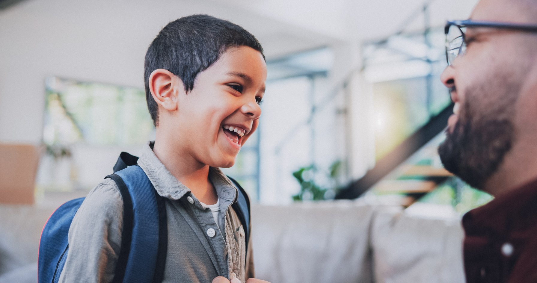 A young boy of Indian ethnicity, wearing a backpack, is laughing heartily while looking at an adult male. The adult, partially visible with glasses and a beard, is also smiling, indicating a warm and happy interaction in an indoor setting.