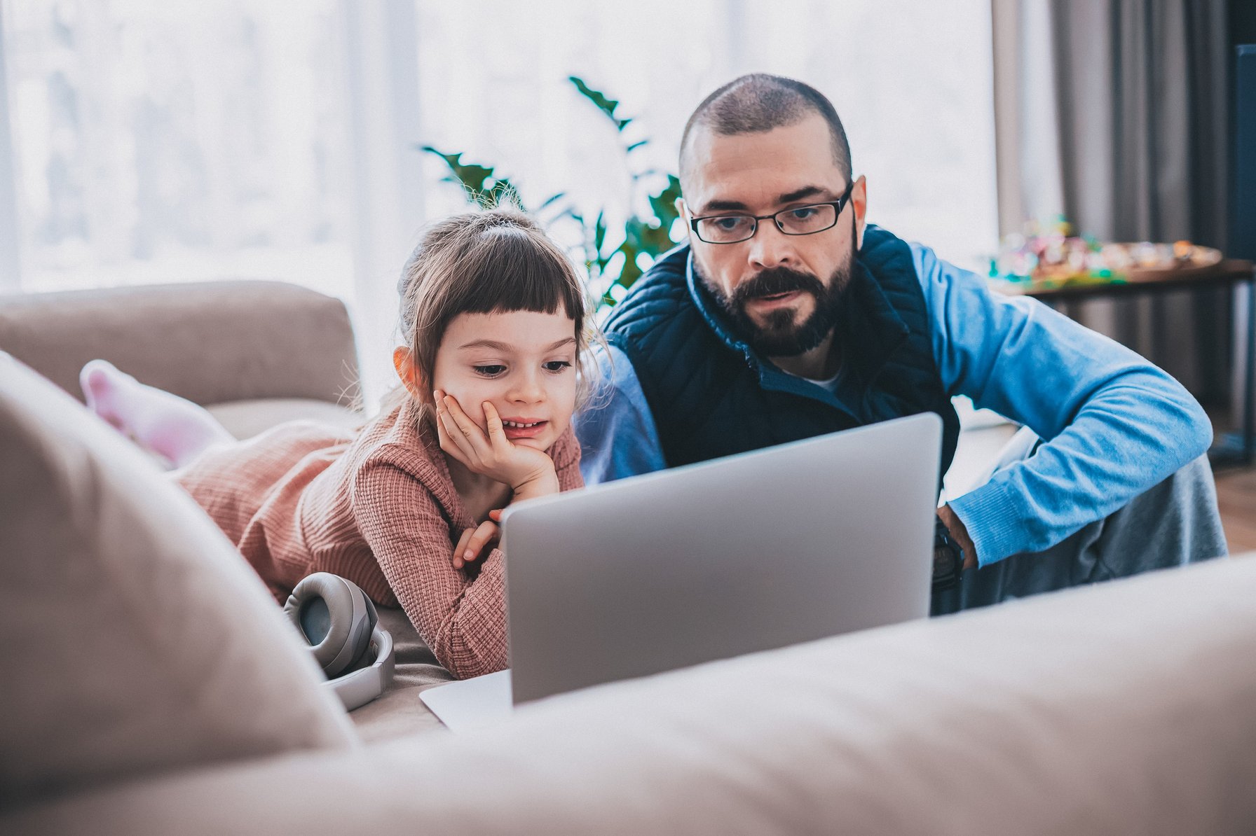 A young girl and an adult man are engaged, looking at a laptop screen together in a cozy living room. The girl lies on a sofa with headphones nearby, while the man sits attentively beside her.