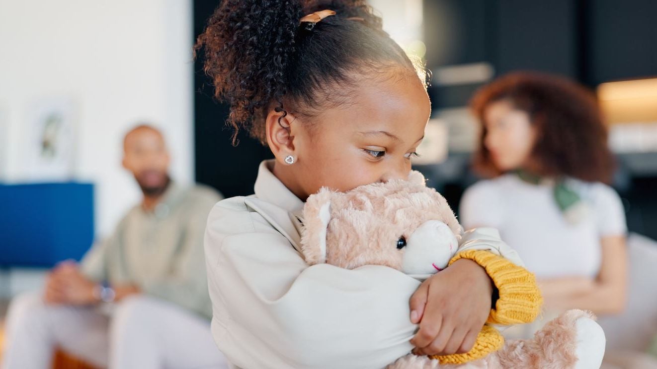 Young girl with anxiety hugging a stuffed animal with parents behind her